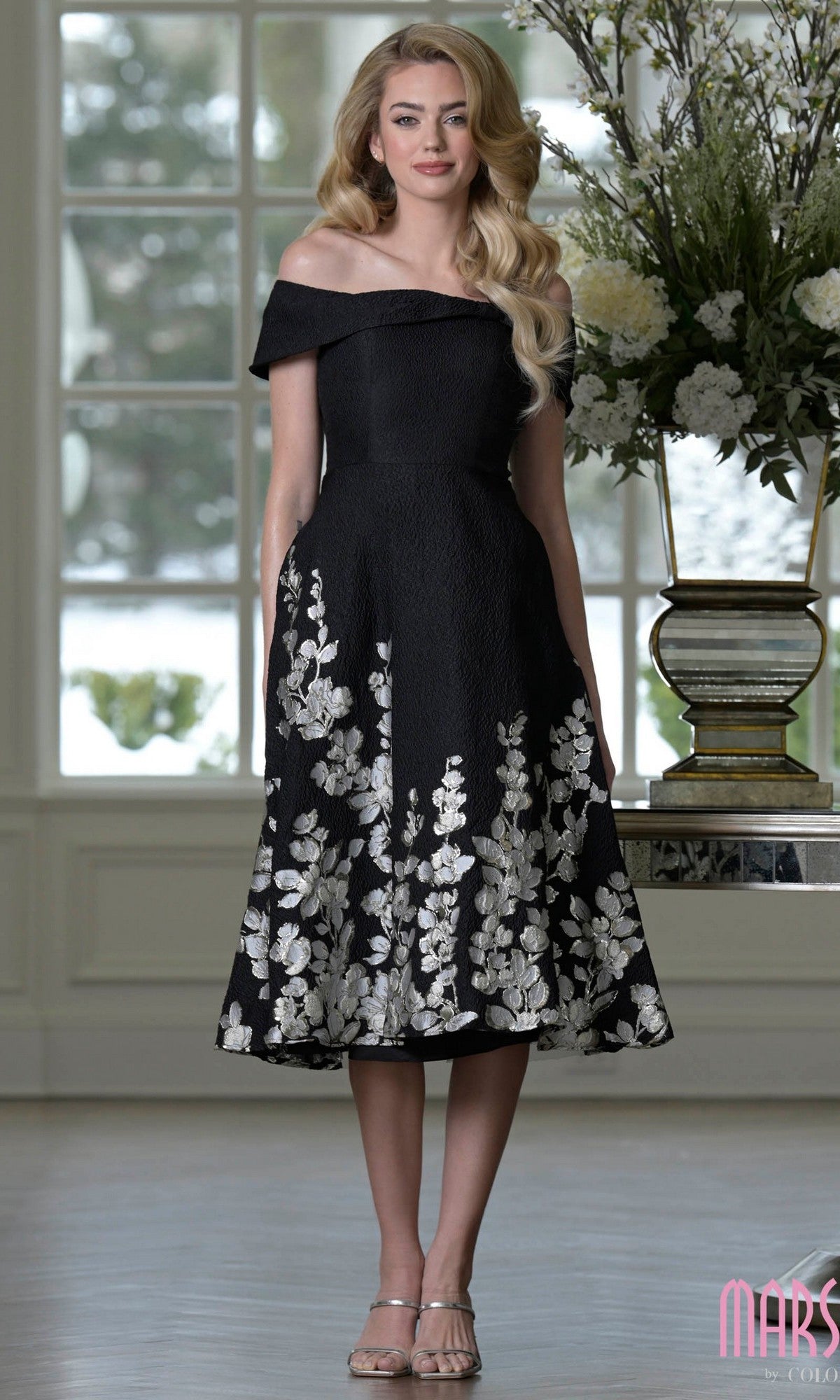 A woman wears the Knee Length Semi-Formal Dress: Marsoni MV1414 in Silver Black, featuring off-the-shoulder styling and floral embroidery, standing indoors by a large window and floral arrangement.